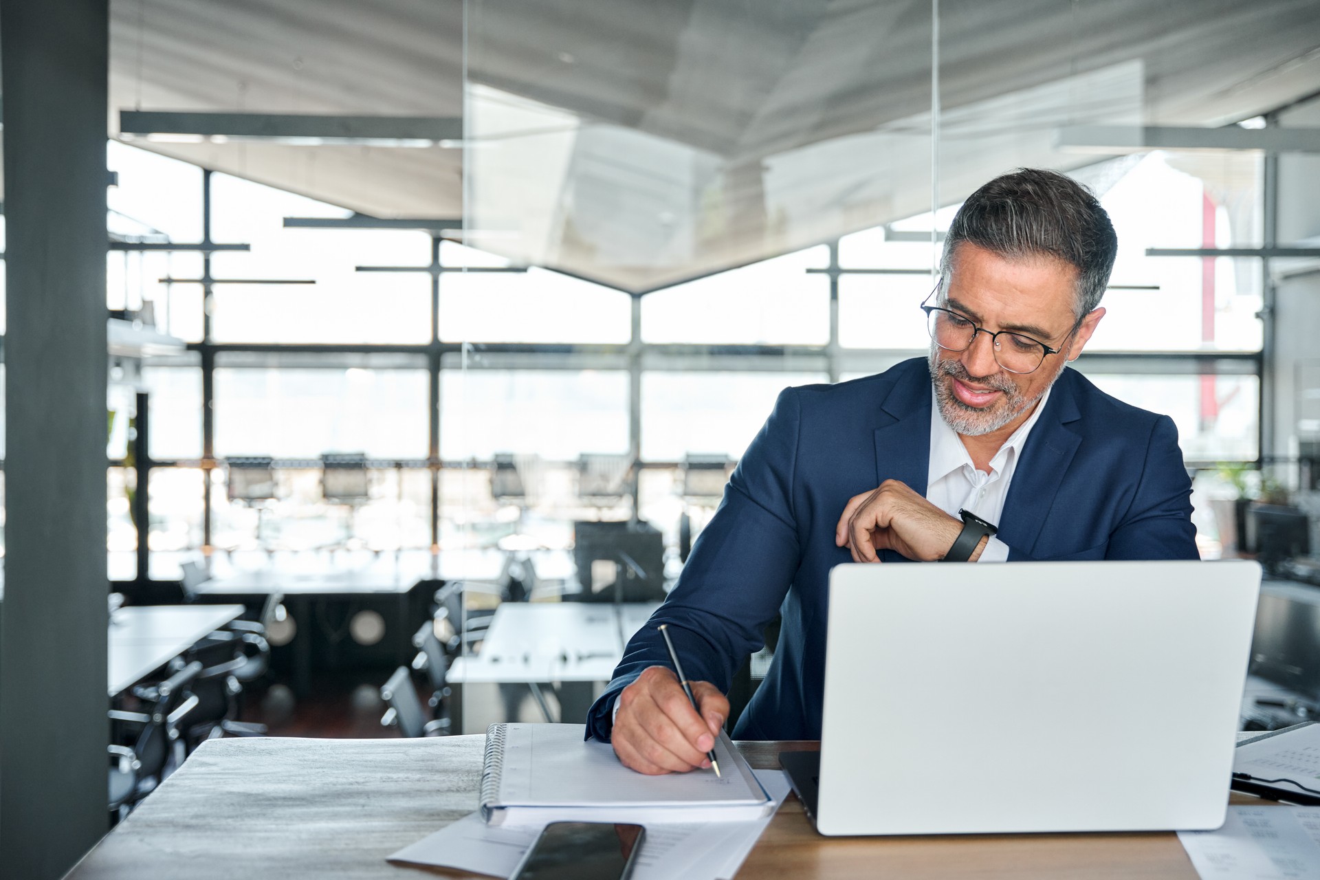 Mid aged business man working on laptop computer in office writing notes.